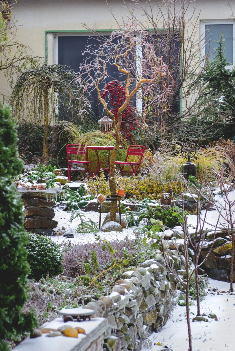 Pod mrazivou krustou uvízl také sníh. Díky němu do popředí vystupují zelené rostliny i zářivě červená barva okrasných jablíček i zahradního nábytku.